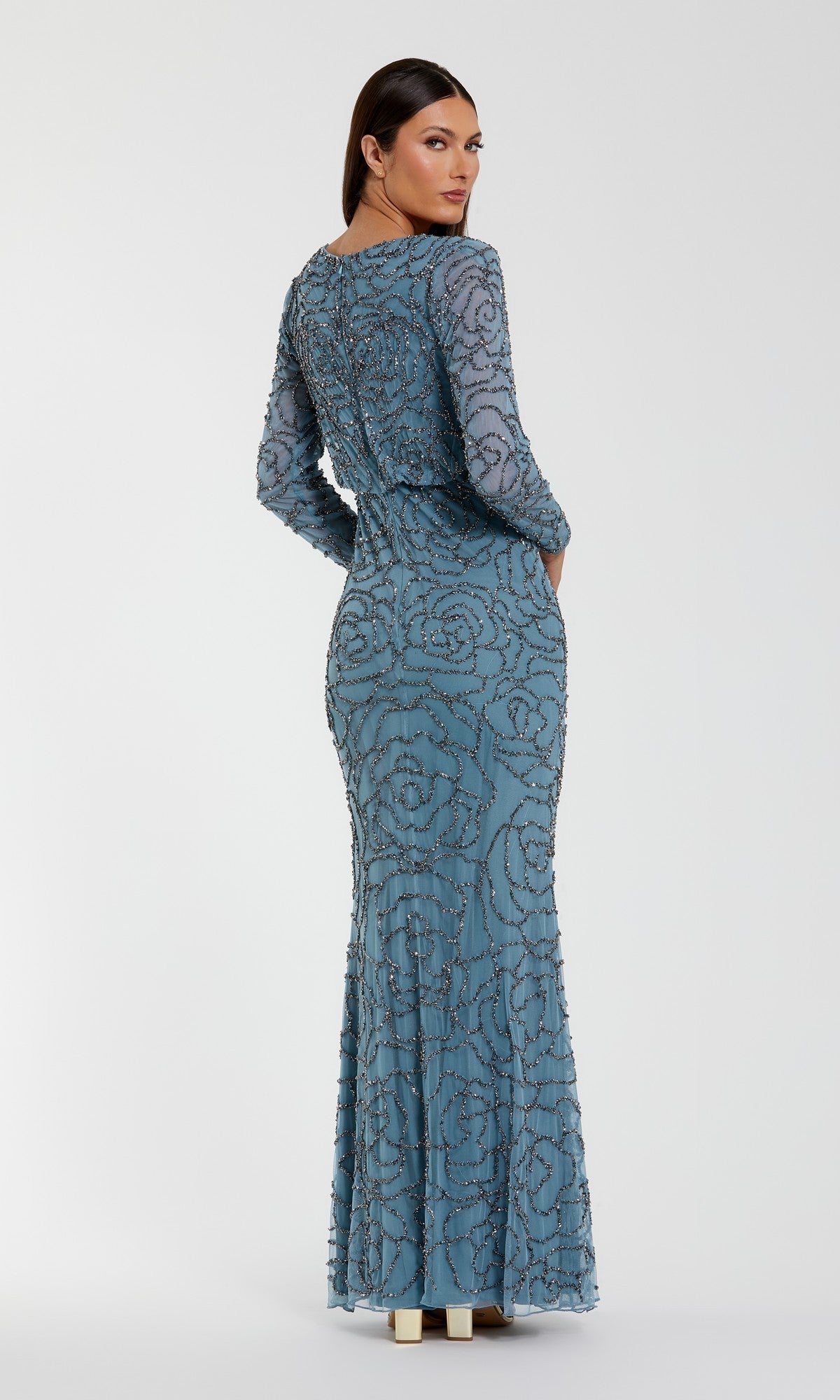 A woman is seen from behind, standing against a plain light background, wearing the Mac Duggal 6189 long formal dress—a fitted blue gown with long sleeves and elegant bead detailing.